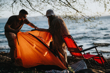 Two friends enjoying outdoor leisure time while setting up an orange tent beside a lake, embodying...