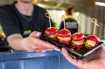 Close-up of a hand holding a tray with four mini burgers, garnished with sesame seeds and fresh...