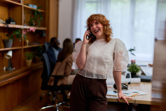 A woman with curly red hair is smiling while talking on the phone in a modern office setting. She is leaning against a desk with papers and a plant on it.