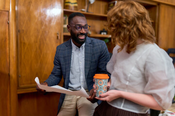 A black man in a suit and a caucasian woman holding a coffee cup are discussing a document in an...