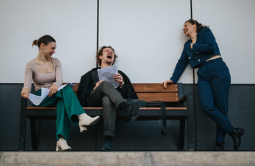 Three colleagues enjoy a light moment on a bench, laughing and relaxing during a break from their...