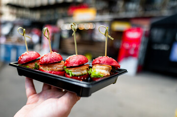 A hand holds four mini burgers with red buns on a black tray, set against a blurred street market background