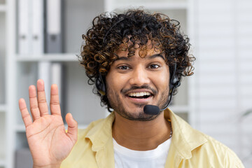 Portrait of a cheerful customer service representative with a headset, smiling and waving at the camera in an office setting.
