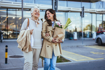 A joyful adult daughter walks with her elderly mother outside a store, both carrying grocery bags...