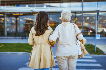 A young woman supports her elderly mother carrying groceries, both clad in light jackets, outside a...