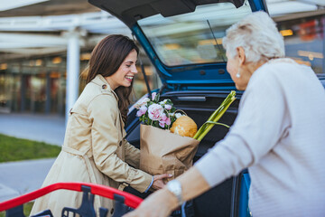 A joyful Caucasian woman assists her elderly mother in transferring grocery bags from a trolley to...