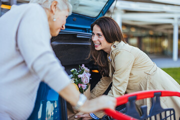 A smiling Caucasian woman assists her elderly mother with groceries, placing bags in the trunk of a...