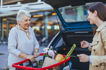 Caucasian adult daughter aids joyful senior mother by loading groceries into the car trunk after...