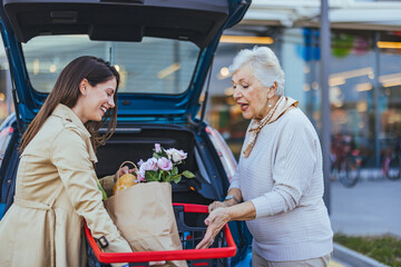 Caucasian woman helps her elderly mother by placing shopping bags into the car trunk outside a...