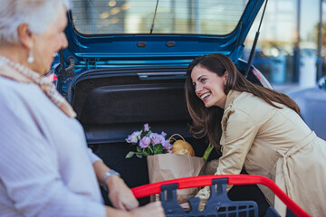A smiling adult daughter assists her elderly mother, loading fresh groceries into the open trunk of...