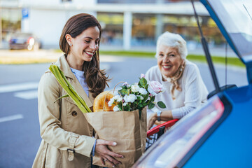 A joyful Caucasian adult daughter places fresh groceries into a car trunk, assisted by her smiling...
