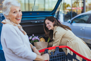 A joyful Caucasian woman assists her elderly mother, loading groceries into a car trunk in a...