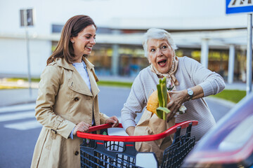 A cheerful Caucasian woman helps her elderly mother stow shopping bags in a car trunk, both enjoy a...