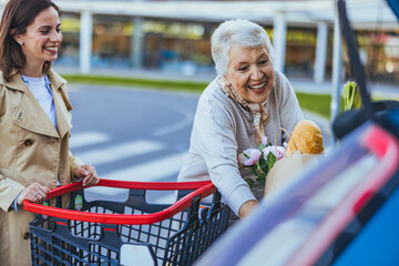 A smiling adult woman assists her joyful senior mother in placing groceries into the car trunk,...