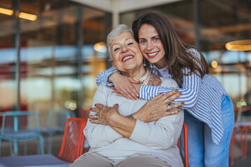 A joyful Caucasian elderly woman and her adult daughter embrace, smiles abound, in a warmly lit...