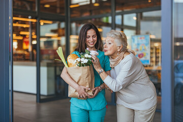 A smiling Caucasian nurse supports an elderly lady holding a bag of groceries outside a store,...