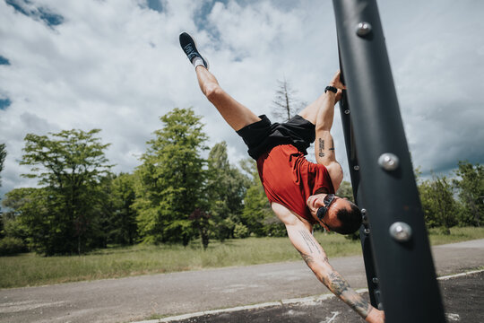 A fit man with tattoos doing a one-arm handstand on a workout bar in a lush green park, showcasing strength and balance under a clear sky.
