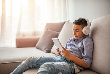 Smiling boy with tablet PC and headphones listening to music or playing game at home. Happy Little...