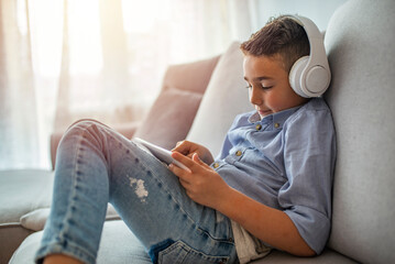 Little boy aged 6 listening to the music using modern tablet. The boy is wearing headphones and...