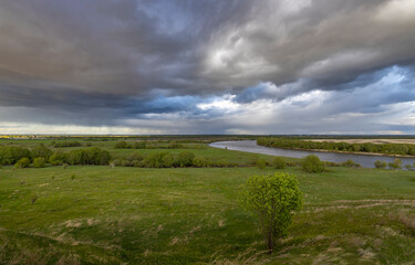 rural landscape with a meandering river cutting through a vast green meadow. The horizon is dominated by a dramatic sky filled with storm clouds, suggesting an impending change in weather.
