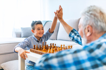 Handsome grandpa and grandson are playing chess and smiling while spending time together at home....