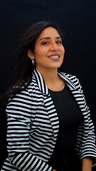chica latina sonriendo con fondo negro usando una blusa negra y una chaqueta de rayas blanca y negra
