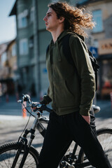 Casual young man with his bicycle in the city, sporting long, wind-blown hair and a relaxed vibe,...