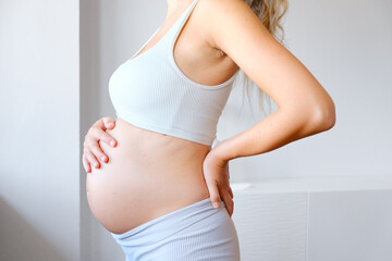 Pregnant Unrecognized Woman Dressed In A Sporty Outfit Holding Her Belly And Standing In A Pose In Profile.  Expecting Child. Home Indoor Interior. Concept Of Pregnancy and New Life.