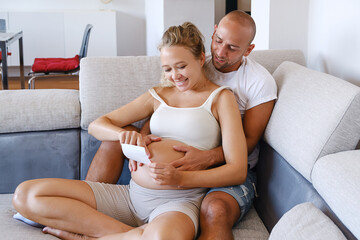 Couple Of  Parents Hold The Scan Of The Baby's Ultrasound, Sitting At Home On The Couch. Concept Of...