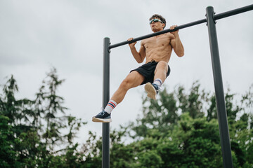 A young athletic man performs pull-ups on metal bars in an outdoor park setting, demonstrating...