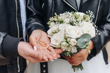 bride and groom in black leather jackets hold wedding rings and bouquet