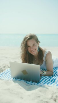 happy modern middle aged woman on the seashore with straw bag, laptop and striped towel.
