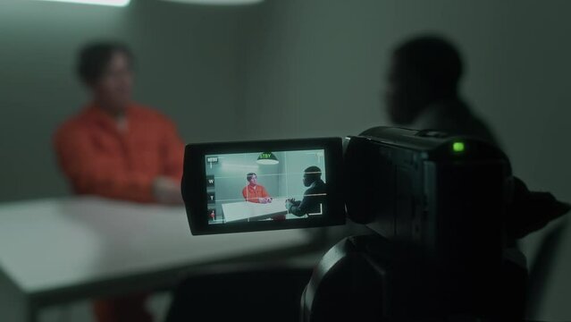 Close-up shot of screen of video camera recording African American law enforcement agent interrogating Caucasian violent crime suspect in orange overalls, in dimly lit room at police station