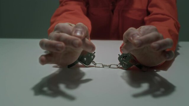Close-up shot of detained crime suspect in handcuffs, orange prison jumpsuit sitting at table in dimly lit interrogation room, twisting fingers, then clasping hands, while being questioned by police