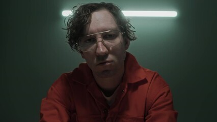 Medium close-up portrait of Caucasian male serial murder suspect with ragged wavy hair, in orange prison jumpsuit sitting in interrogation room or jail cell, looking at camera, while being questioned
