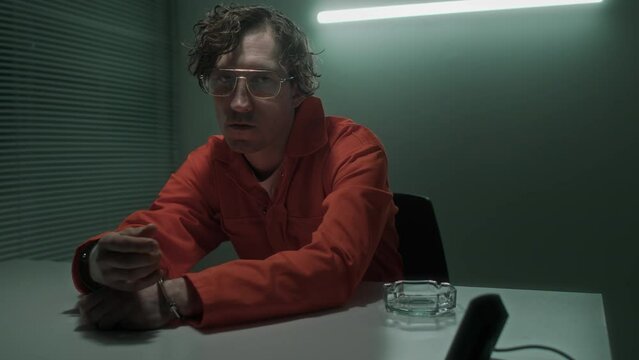 Medium portrait of Caucasian male serial murder or terror crime suspect with dirty wavy hair, glasses, in orange jumpsuit sitting in interrogation room, fidgeting with handcuffs, looking at camera