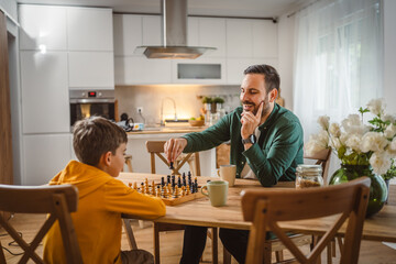 Father with cup of hot drink and son play chess together at home