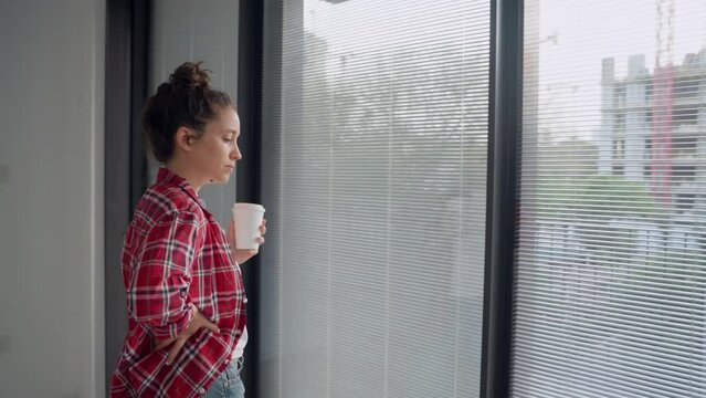 Woman looking outside big windows with her small dog standing in empty apartment. Female home owner is sad to leave her place or have her parter leave. Small dog walking around. Card boxes on floor
