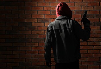 Thief in hoodie with gun against red brick wall, back view. Space for text