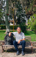 young latin man with serious expression sitting looking aside with travel backpack on a sunny day