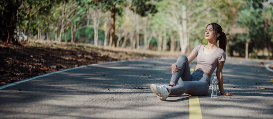 Young Woman Resting on a Road in a Park During a Workout, Wearing Athletic Clothing, with a Water...