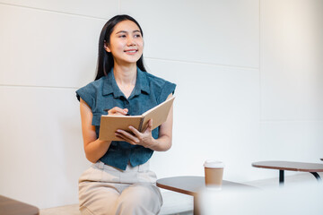 Young Woman Smiling While Writing in Notebook at Modern Cafe with Coffee Cup on Table, Casual...