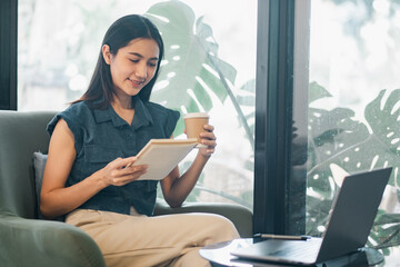 Young Woman Enjoying Coffee While Working on Digital Tablet at Home Office with Laptop and Green...