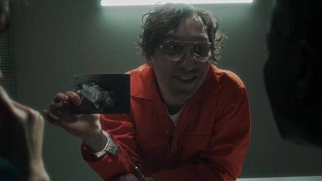 Medium shot of detectives interrogating psychotic murder suspect in orange jumpsuit, who is taunting them with maniac laughter, shaking photo of victim body, shouting, then lighting cigarette