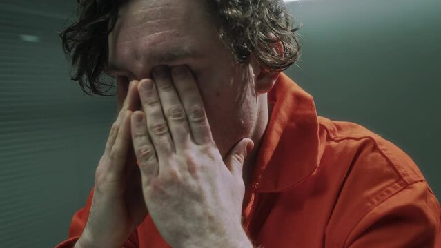 Close-up shot of young Caucasian male serial murder suspect in orange prison jumpsuit sitting in interrogation room in pretrial detention facility, rubbing eyes while considering crime confession