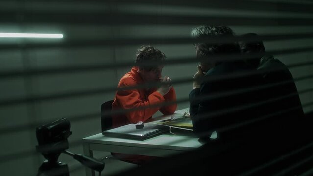 Full shot through window blinds of diverse male and female federal agents questioning murder suspect in orange prison robe in detention facility, filming on video camera, man confessing to crime