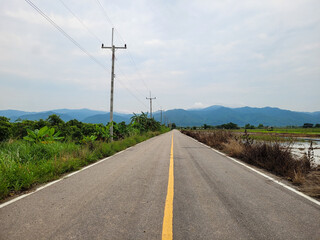 Fototapeta premium A long, straight road stretches into the distance, flanked by lush greenery and power lines, leading towards a range of misty mountains under a cloudy sky. This image captures the serene and expansive