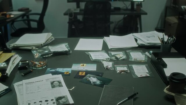 Close-up no people shot of table with criminal profile bulletins, homicide scene photos, evidence envelopes, clues in plastic bags, ashtray, cigarettes, in situation room at busy police department