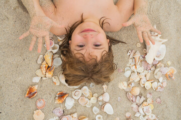 Kid laying on sandy beach with Shells. Summer kids. Portrait from above of a boy holding seashell...