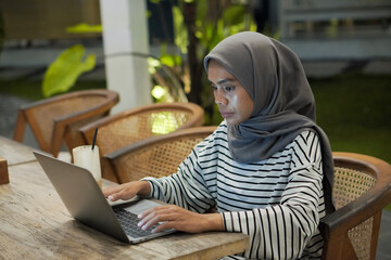 A muslim woman in hijab uses a laptop on a wooden table in an outdoor cafe at night